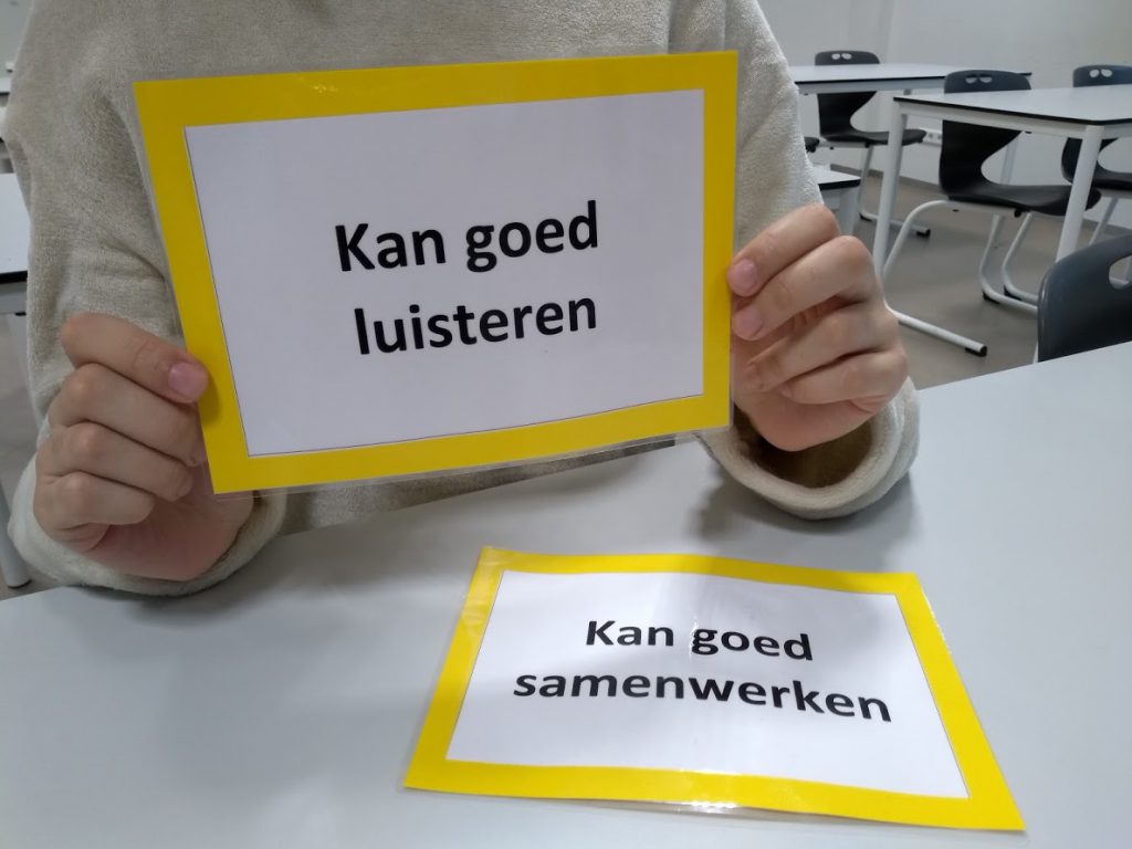 Leerling houdt het complimentenkaartje vast waarop 'Kan goed luisteren' staat. Op de tafel ligt het kaartje 'Kan goed samenwerken'.